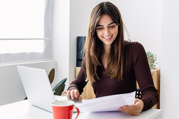 Young smiling pretty adult woman working on laptop while checking home invoices or financial...
