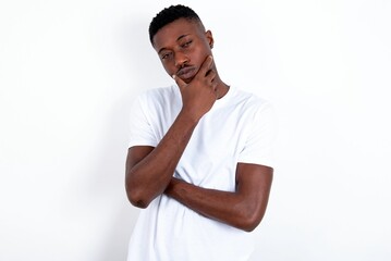 Thoughtful smiling young handsome man wearing white T-shirt over white background  keeps hand under chin, looks directly at camera, listens something with interest. Youth concept.