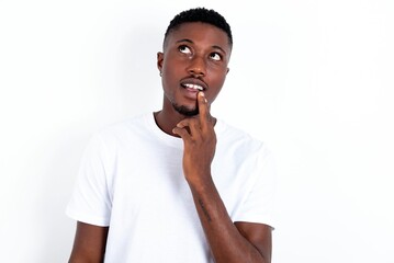 young handsome man wearing white T-shirt over white background with thoughtful expression, looks to the camera, keeps hand near face, bitting a finger thinks about something pleasant.