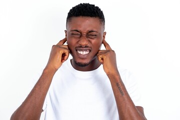 Stop making this annoying sound! Unhappy stressed out young handsome man wearing white T-shirt over white background making worry face, plugging ears with fingers, irritated with loud noise.