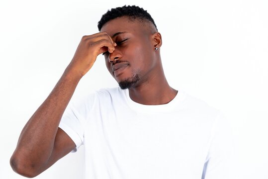 Very upset, young handsome man wearing white T-shirt over white background touching nose between closed eyes, wants to cry, having stressful relationship or having troubles with work
