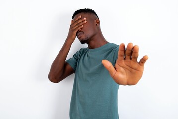 young handsome man wearing green T-shirt over white background covers eyes with palm and doing stop gesture, tries to hide. Don't look at me, I don't want to see, feels ashamed or scared. - obrazy, fototapety, plakaty