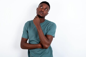 Face expressions and emotions. Thoughtful young handsome man wearing green T-shirt over white background holding hand under his head, having doubtful look.