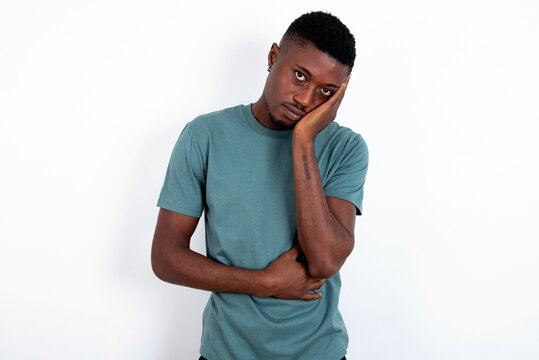 Very bored young handsome man wearing green T-shirt over white background holding hand on cheek while support it with another crossed hand, looking tired and sick.