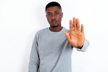 young handsome man wearing grey sweater over white background shows stop sign prohibition symbol keeps palm forward to camera with strict expression