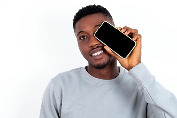 young handsome man wearing grey sweater over white background holding modern smartphone covering one eye while smiling
