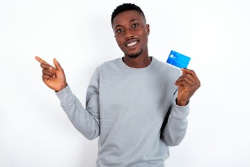 Smiling young handsome man wearing grey sweater over white background showing debit card pointing finger empty space