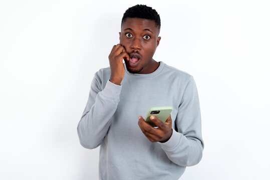 Afraid funny young handsome man wearing grey sweater over white background holding telephone and bitting nails
