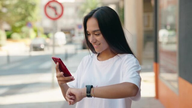 Young beautiful hispanic woman using smartphone looking watch at street
