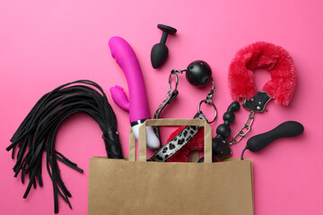 Paper shopping bag with different sex toys on pink background, flat lay