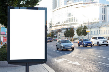 White blank vertical billboard in the city. Bright sun reflects off the office building. Cars drive along the highway. Mock-up.