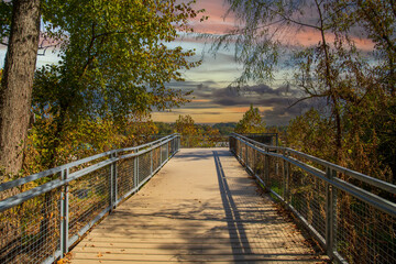 Fototapeta na wymiar a gorgeous autumn landscape at Lakeshore Park with wooden footpath to a deck surrounded by a gray metal hand rail, autumn colored trees and lush green trees and grass with powerful clouds at sunset