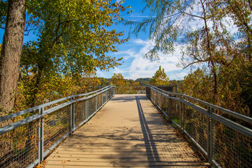 a gorgeous autumn landscape at Lakeshore Park with wooden footpath to a deck surrounded by a gray metal hand rail, autumn colored trees and lush green trees and grass with blue sky and clouds - obrazy, fototapety, plakaty