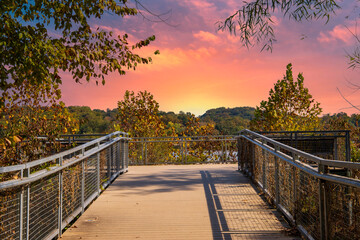 Plakat a gorgeous autumn landscape at Lakeshore Park with wooden footpath to a deck surrounded by a gray metal hand rail, autumn colored trees and lush green trees and grass with powerful clouds at sunset