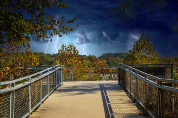 Obraz na płótnie Canvas a gorgeous autumn landscape at Lakeshore Park with wooden footpath to a deck surrounded by a gray metal hand rail, autumn colored trees and lush green trees and grass with storm clouds and lightning
