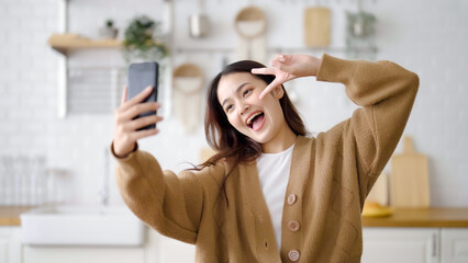 Pretty young asian female smile and stand at kitchen room. She having fun taking selfie and video call talking with her friend