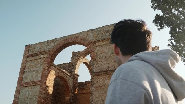 Young man stops in front of an old ruined chapel architecture from a distance and walks towards it. Spain.