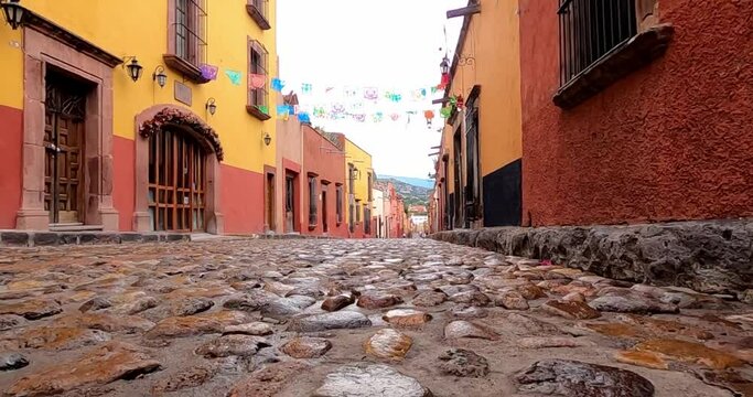A vibrant cobblestone street in a Mexican town, lined with colorful colonial buildings. Papel picado decorations hang across the street, creating a festive atmosphere under a bright sky.
