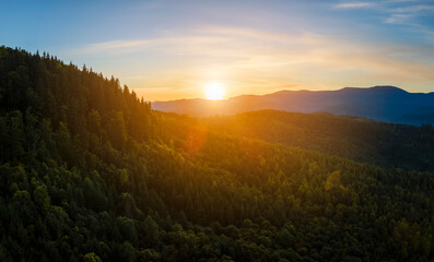 Aerial view of bright foggy morning over dark hills with mountain forest trees at autumn sunrise. Beautiful scenery of wild woodland at dawn