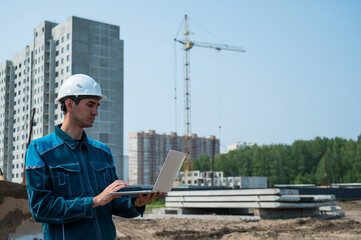 A male builder in a helmet stands against the background of a construction site with a laptop in his hands. 
