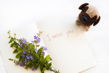 i miss you message handwriting on notebook with dog toy purple flowers arrangement flat lay postcard style on background wooden