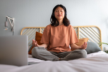 Front view of teen asian girl meditating at home sitting on bed following online meditation,...