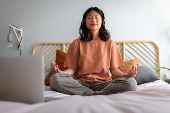 Front view of teen asian girl meditating at home sitting on bed following online meditation, listening with headphones.