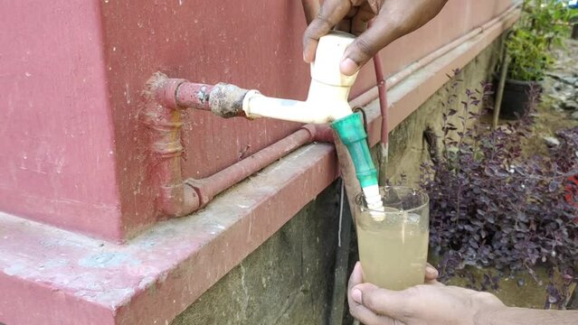 Hand holding and filling glass with less contaminated dirty, rusty and muddy brown drinking water from outdoor wall tap pipe. Indian and African water scarcity, shortage, crisis or impurity concept.