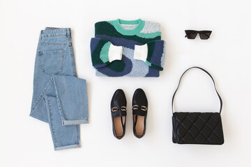 Blue jeans, blue knitted sweater, black loafers or flat shoes, bag and sunglasses lying on white background. Overhead view of women's casual outfit. Trendy stylish women clothes. Flat lay, top view.