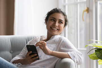 Peaceful Indian woman relaxing on cozy chair with smartphone, smile staring aside enjoy carefree...