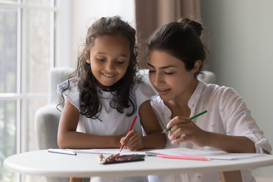 Attractive young Indian mother and little daughter drawing pictures in paper with colored pencils. Loving family enjoy communication and common creative hobby spend leisure at home. Artistic pastime