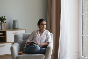 Attractive pensive Indian woman resting on cozy armchair staring into distance spend weekend...
