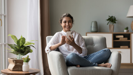 Young smiling Indian woman sits on armchair holding modern cellphone in hands looking at camera. Happy female enjoy distancing communication, share sms and messages, calling using smartphone gadget