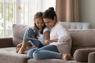 Little cute Indian girl and mom use digital tablet, resting together at home, buying goods through...