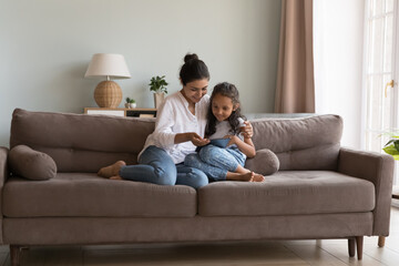Indian woman and little daughter spend time using digital tablet, sit together on couch at home, buying goods, purchasing toys through retail webstore enjoy new application for fun or kid development