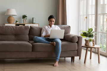 Serious Indian woman put wireless computer on laps texting e-mail to client sits on cozy couch in...