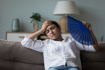 Indian woman suffers from heat inside, cools herself waving blue paper fan, leaned on sofa in...