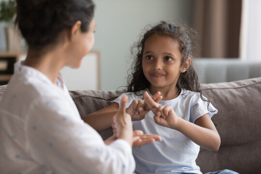 Deaf Indian preschooler girl and young mother showing symbols with hands using visual-manual gestures enjoy communication seated on couch at home. Hearing loss and disability, sign language usage
