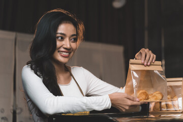 portraits of beautiful waitress pick a croissant from customer order and pack in bakery bag and present bakery to customer. the happiness woman in bakery small business on waitress uniform.