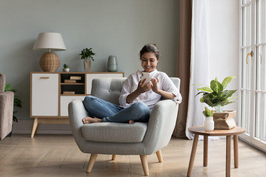 Beautiful Indian woman relaxing on armchair in fashionable cozy living room use smartphone, chatting on-line, make call or order via electronic services. Modern tech usage for fun and communication