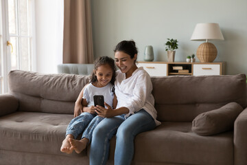 Obraz premium Attractive young Indian woman her little daughter having fun together using modern smartphone, staring at cellphone screen spend leisure on internet enjoy on-line game, new mobile application at home