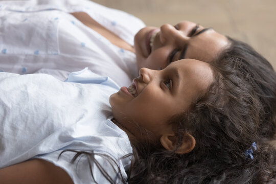 Cute little Indian girl and loving smiling mother lying together on floor indoors, close up. Young woman spend leisure with preschooler daughter, enjoy carefree time and happy motherhood. Love, bond