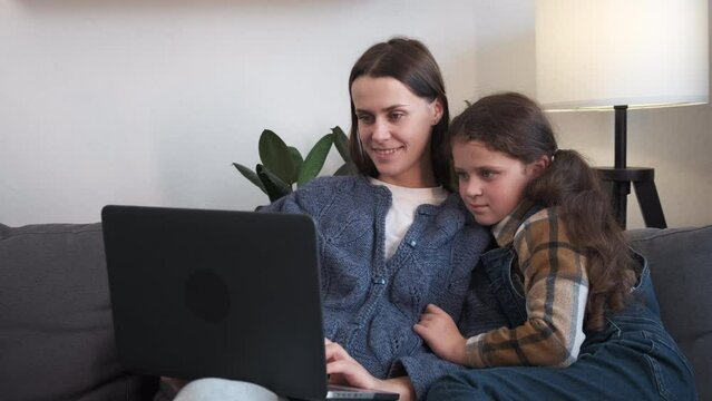 Smiling beautiful young mother teaching adorable little daughter to use laptop sit on comfy couch in living room at home, happy mum and pretty girl having fun, watching video, browsing apps concept
