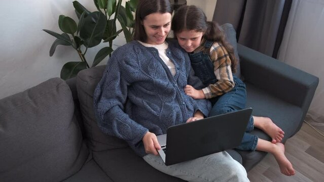 Relaxed young mother with adorable little daughter using laptop, happy mum and girl kid sitting on cozy couch in living room at home, watching cartoons online or play video game, family having fun