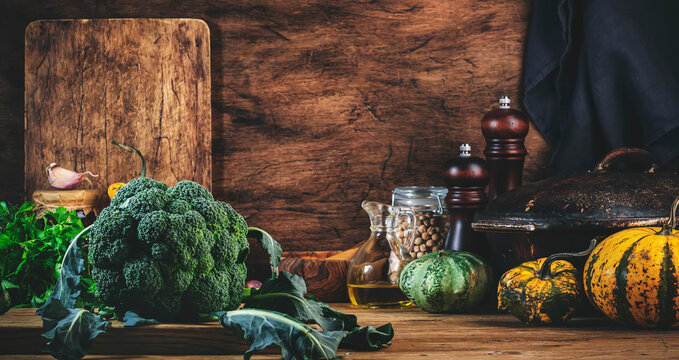 Autumn food cooking background with organic farm broccoli cabbage, pumpkins, herbs and spices on rustic wood kitchen table with cast iron pan, spice grinders, cutting board. Banner