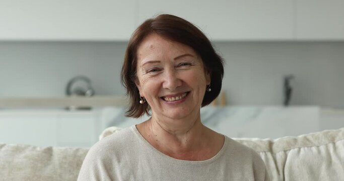 Head shot of mature pretty woman having pleasant smile and attractive appearance pose seated on sofa in cozy living room at home alone. Boomer generation female portrait, carefree retired life concept