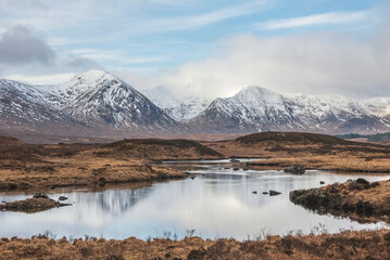 Majestic Winter panorama landscape image of mountain range and peaks viewed from Loch Ba in Scottish Highlands with dramatic clouds overhead