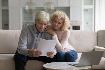 Concerned older couple, wife and husband sit on sofa at home read received formal notification from bank about mortgage denial, unpaid taxes, financial issues, learn insurance terms looking serious