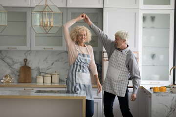 Fototapeta na wymiar Attractive mature wife and older husband in aprons holding hands swirling having fun distracted from cooking in fashionable kitchen, moving to music, dancing together at home feel carefree look happy