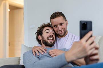 Obraz premium Cute young gay couple video calling their friends in their living room at home. Two male lovers smiling cheerfully while greeting their friends on a smartphone. Young gay couple sitting together.